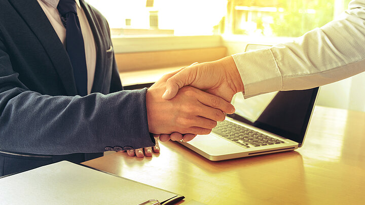 Zdjęcie uścisku rąk nad biurkiem. Na blacie widoczny laptop i notatnik.