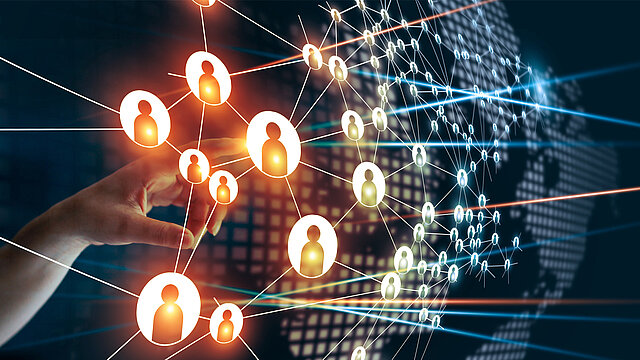 Abstrakcyjna grafika z piktogramami osób powiązanych ze soba promoenistymi liniami. W tle ludzka dłoń dotyka jednego z piktogramów.